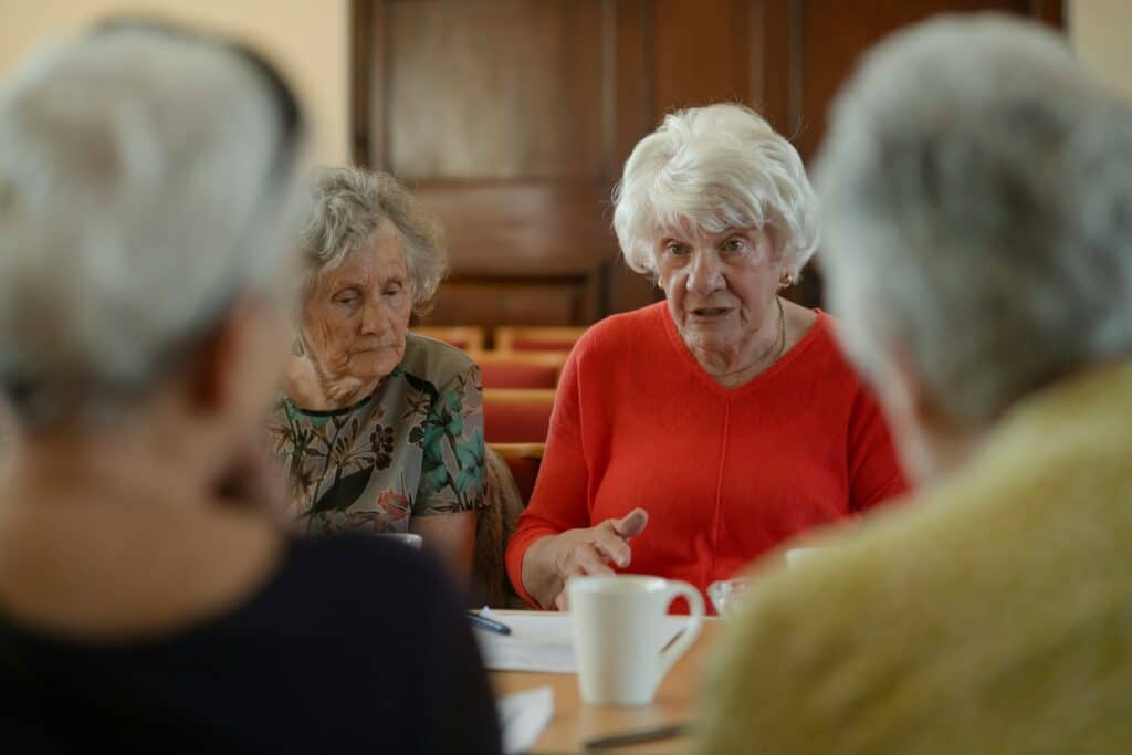 Elderly women sit around a table, talking.