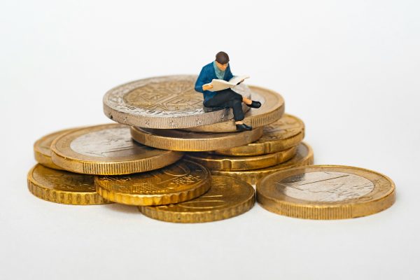 Miniature person sitting on stack of coins reading newspaper